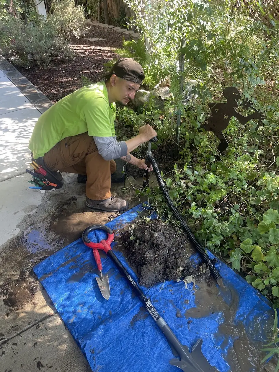 Technician fixing a broken sprinkler pipe in a planter next to a driveway, on a tarp to keep things clean