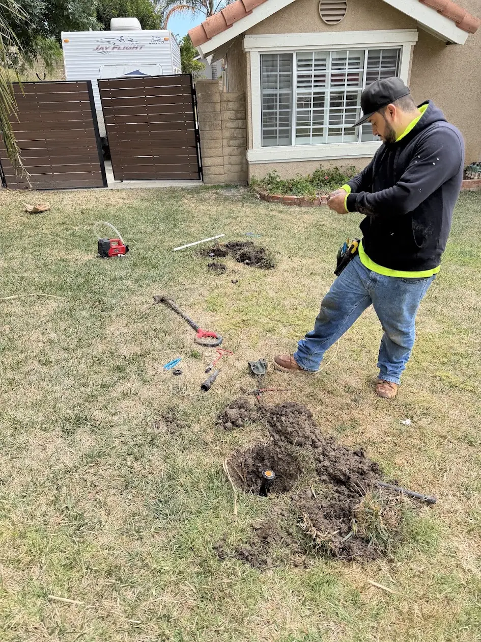 Technician replacing a broken sprinkler head in a front lawn, with tools and the dug-out head beside him