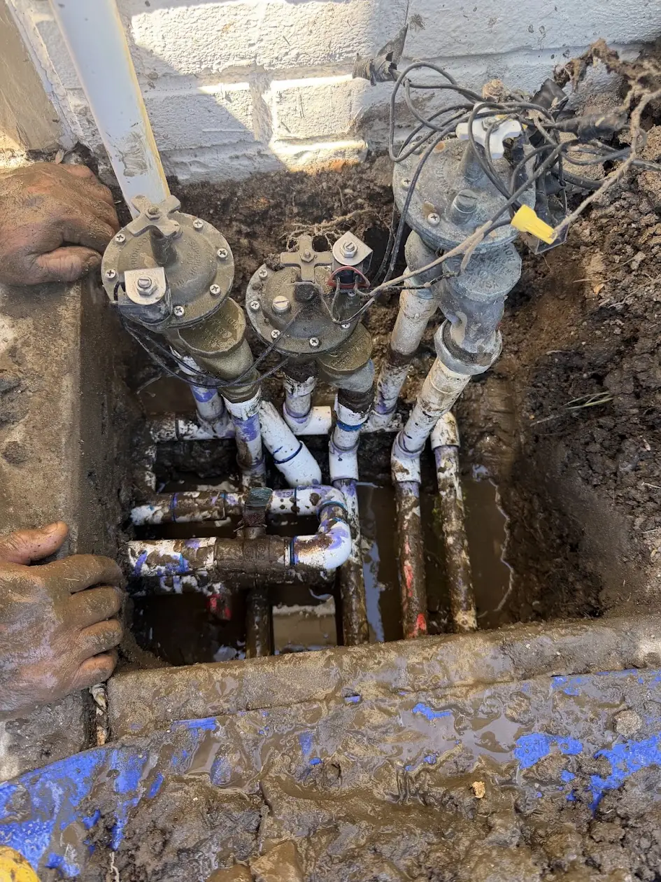 Sprinkler valve manifold being repaired in a muddy trench after an underground leak
